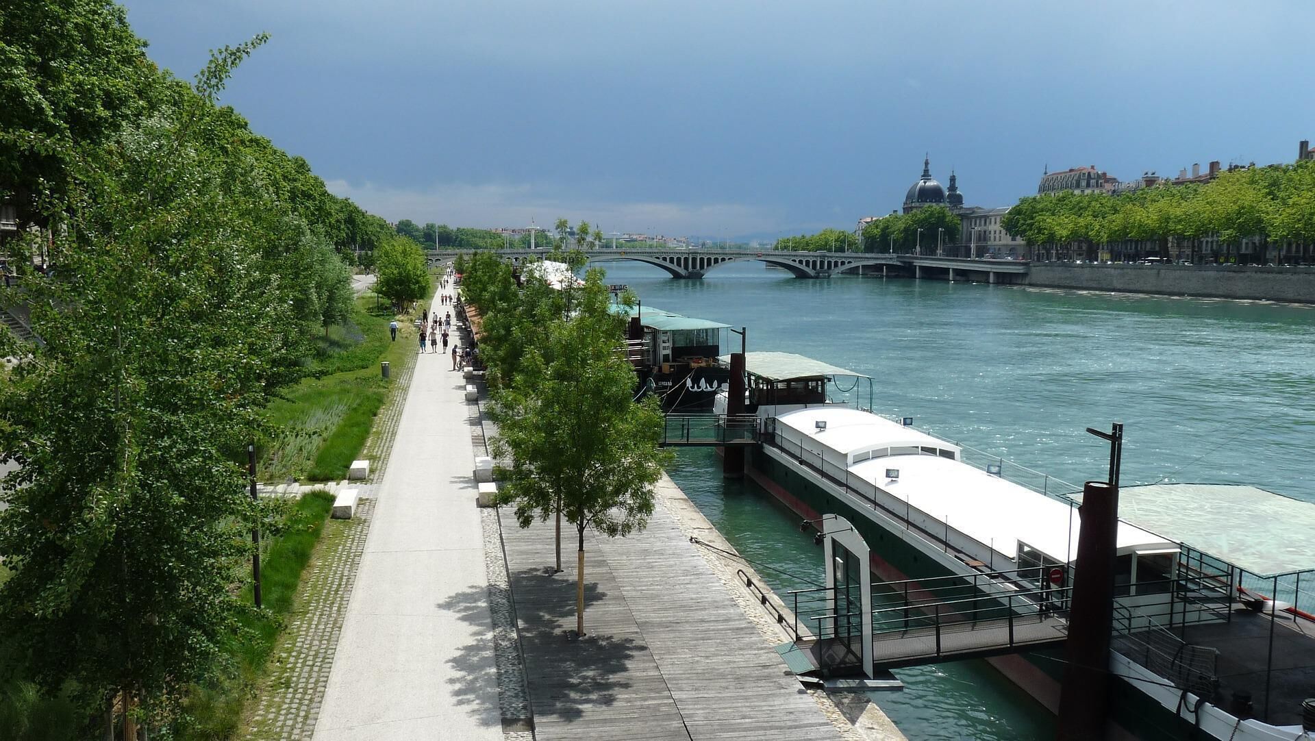 A riverside walkway with a bridge in the background, a boat docked on the river, and a row of trees along the path.