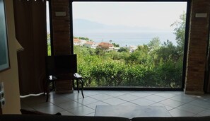 Ein Zimmer mit einer großen Schiebeglasdoor, die auf einen Balkon mit Blick auf das Meer und die grüne Umgebung führt.