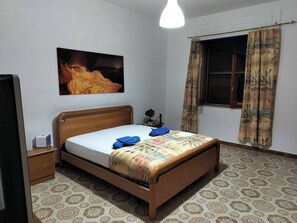 Une chambre avec un lit en bois, une table de chevet, un téléviseur fixé au mur, un sol à motifs et une fenêtre avec des rideaux.