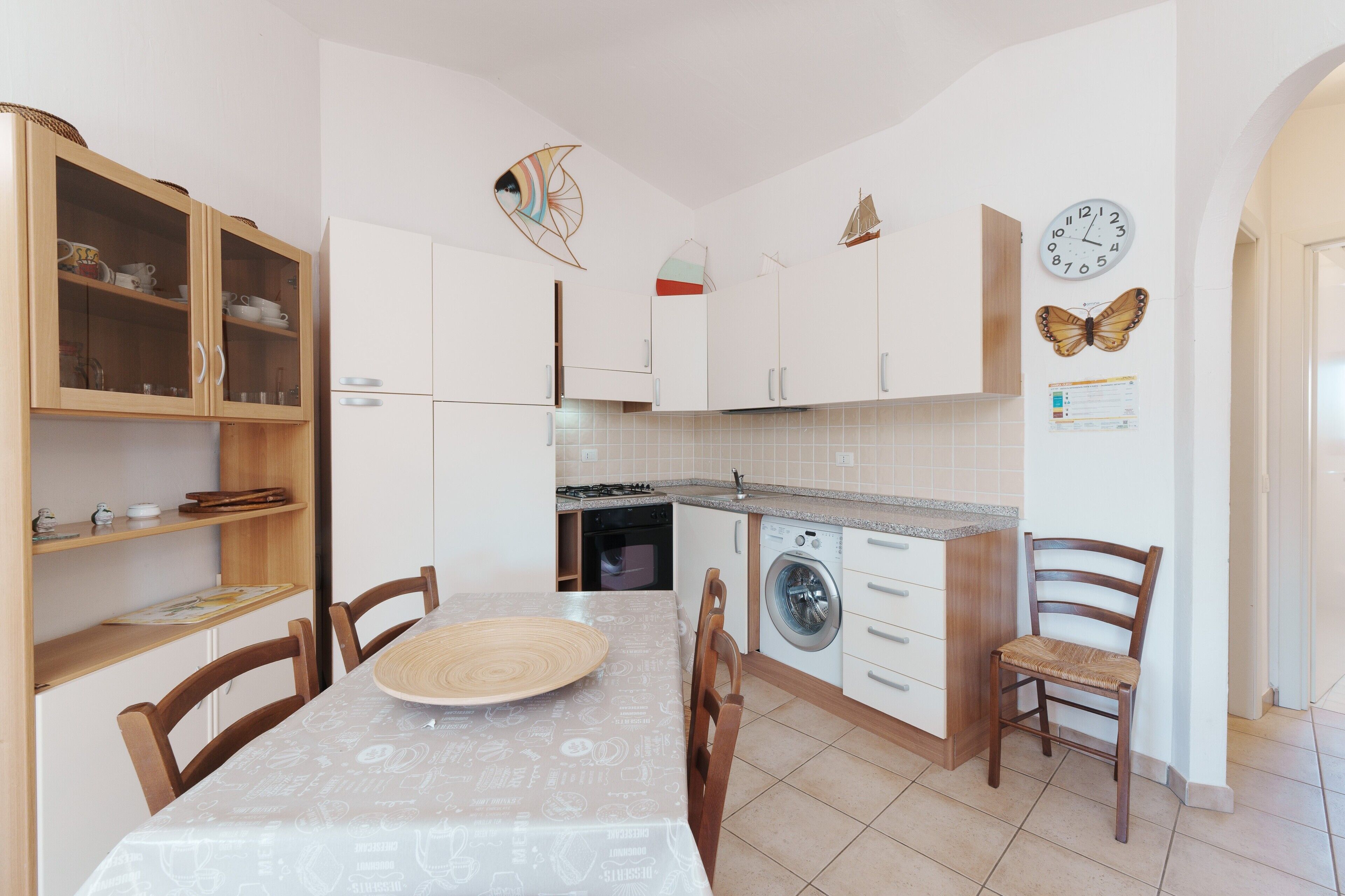 A kitchen with a dining table, chairs, cabinets, a washing machine, and a clock on the wall.