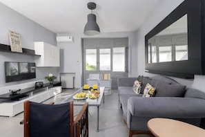 Un salon moderne avec un canapé gris, une table à manger avec des chaises, un téléviseur et une grande fenêtre.