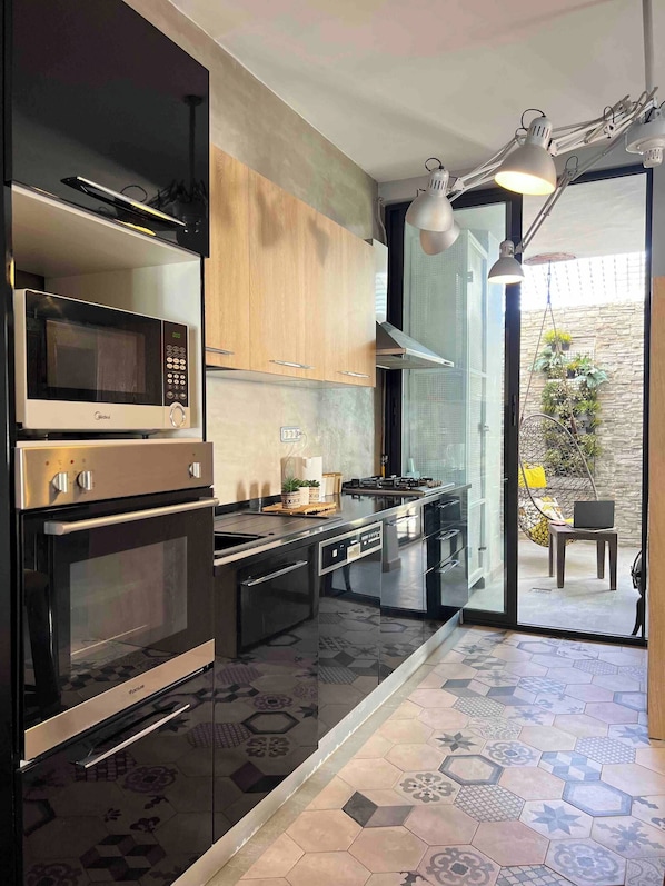 Une cuisine moderne équipée d’un four à micro-ondes, d’un four traditionnel et d’une cuisinière, avec des meubles en bois et une vue sur un espace extérieur agrémenté de plantes et d’un fauteuil suspendu.