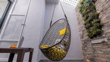 Une chaise suspendue avec des coussins jaunes, une table en osier avec une bougie et un mur de pierre agrémenté de plantes en pot.