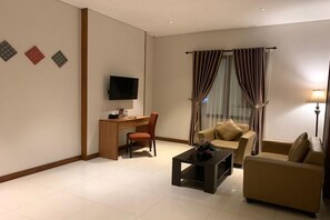 A modern living room with a flat-screen TV, a wooden desk, a brown sofa, a coffee table, and a window with curtains. - Sany Rosa Hotel (Bandung)