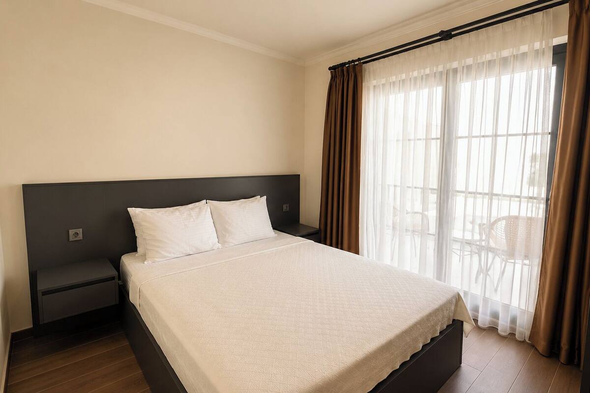 A neatly made bed in a hotel room with a large window, a bedside table, and a dark headboard.