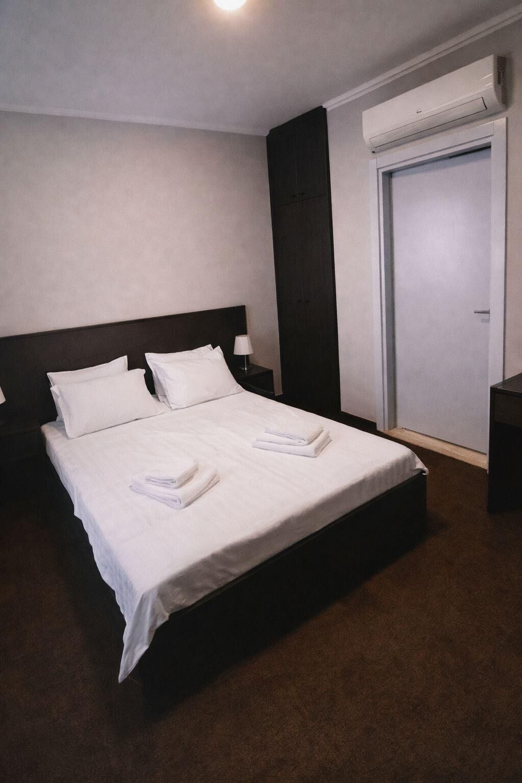A neatly made bed with white linens and towels, a dark headboard, a nightstand with a lamp, and a closed door.