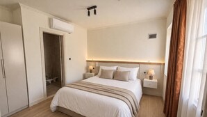 A hotel room with a bed, bedside tables, an air conditioning unit, and a door leading to a bathroom.