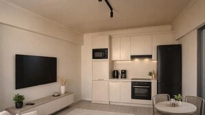 A modern living room with a sofa, a dining table with chairs, a kitchen area with appliances, and a flat-screen TV mounted on the wall.
