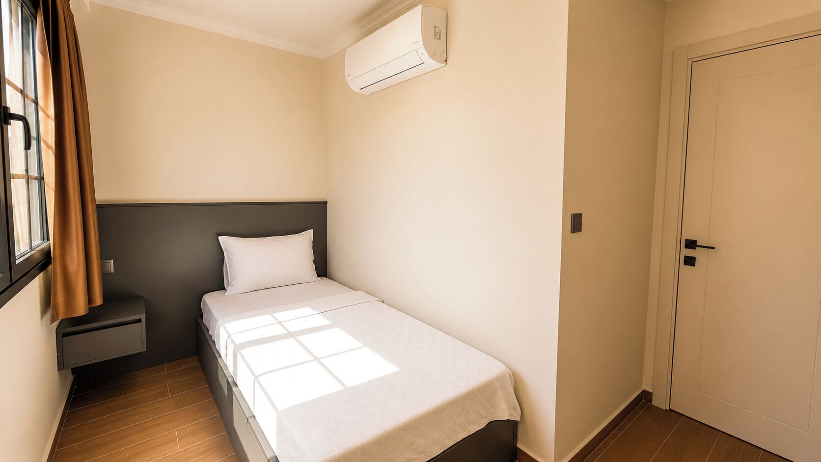 A small, single-bed room with a window, a bedside table, a nightstand, and a door.