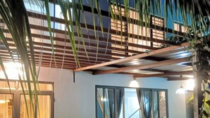 Un área de estar al aire libre con mesa y bancos, rodeada de plantas y una estructura cubierta.