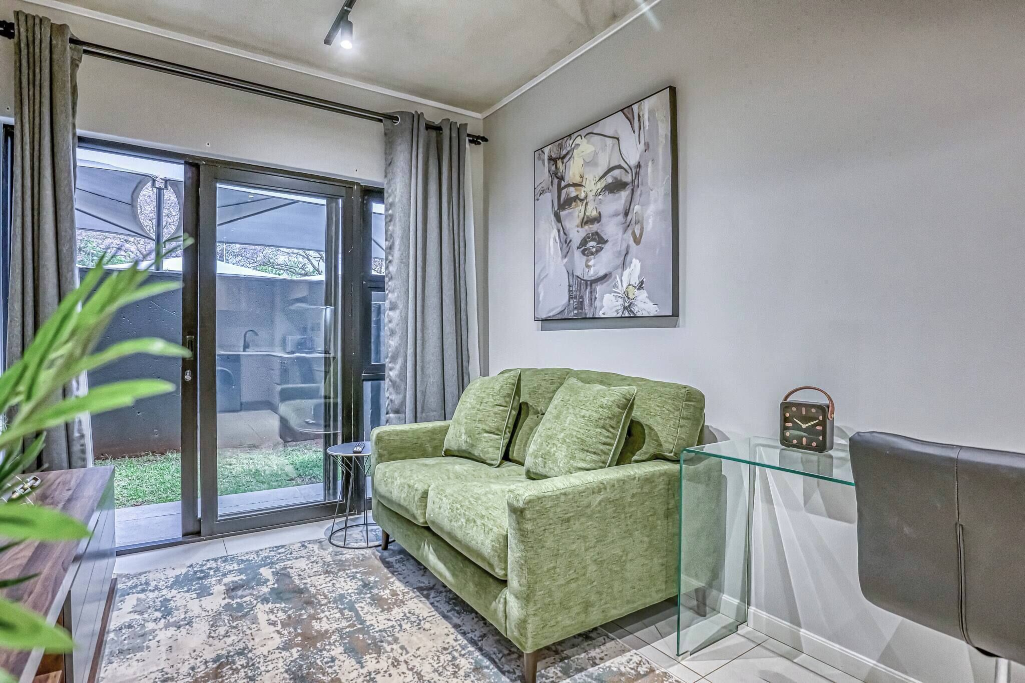 Ein Wohnzimmer mit einem grünen Sofa, einem Glastisch, einer Wanduhr und einer Glastür, die zu einem Außenbereich führt.