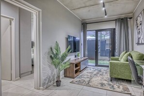 Ein Wohnzimmer mit Fernseher, einer Couch, einer Pflanze und einer Glastür, die auf eine Terrasse führt.