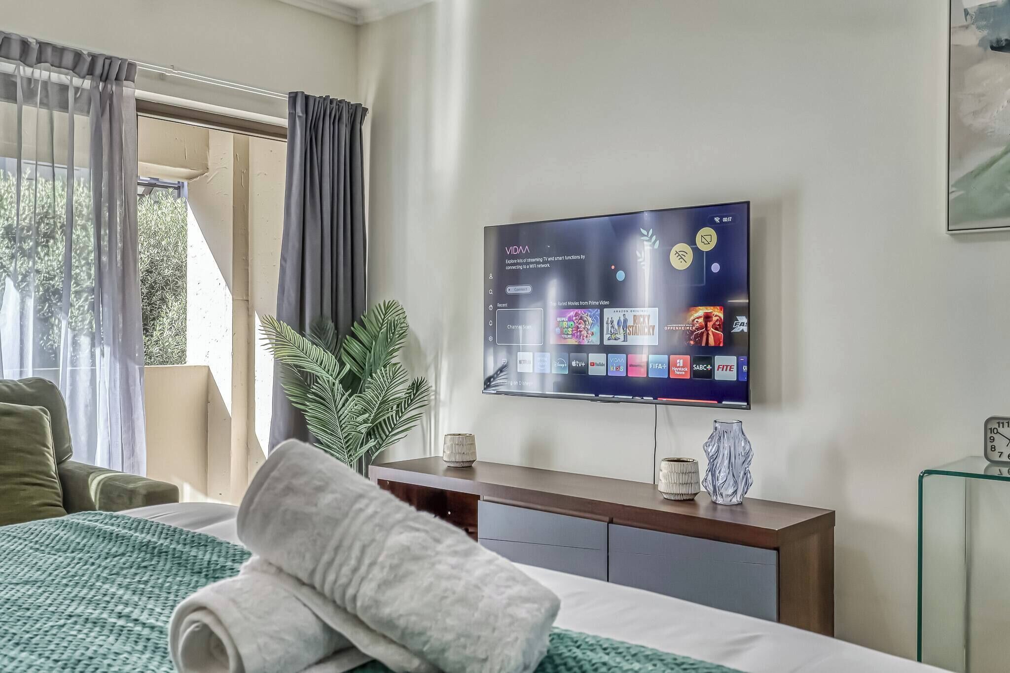Ein Hotelzimmer mit einem Bett, einem an der Wand befestigten Fernseher, der Streaming-Dienste anzeigt, einem Nachttisch mit einer Vase und einem Fenster mit Vorhängen.