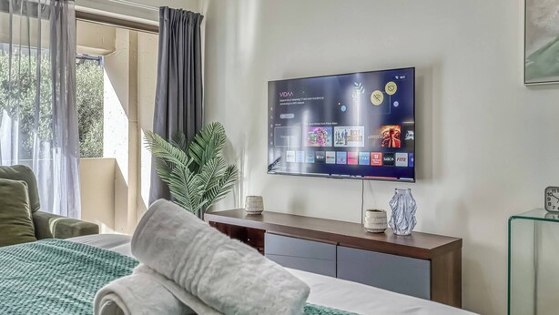 Una camera d'albergo con un letto, una televisione a muro che mostra servizi di streaming, un comodino con un vaso e una finestra con tende.