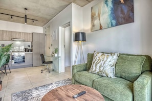 Ein modernes Wohnzimmer mit einem grünen Sofa, einem Couchtisch, einer Fernbedienung, einer Küchenzeile und einer Stehlampe.