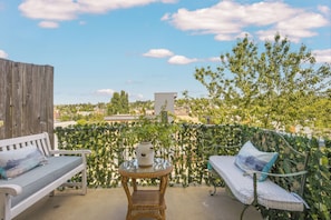 Ein Balkon mit einem Korbstuhl, zwei weißen Sitzbänken und Blick auf eine Stadtlandschaft.