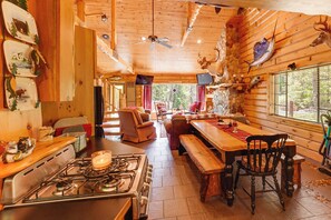 Interior rústico de una cabaña con techo de madera, una mesa de comedor, una estufa y un rincón de estar acogedor.