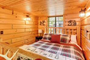 Interior de una cabaña de madera con una cama, una mesita de noche, una ventana con vistas y un reloj en la pared.