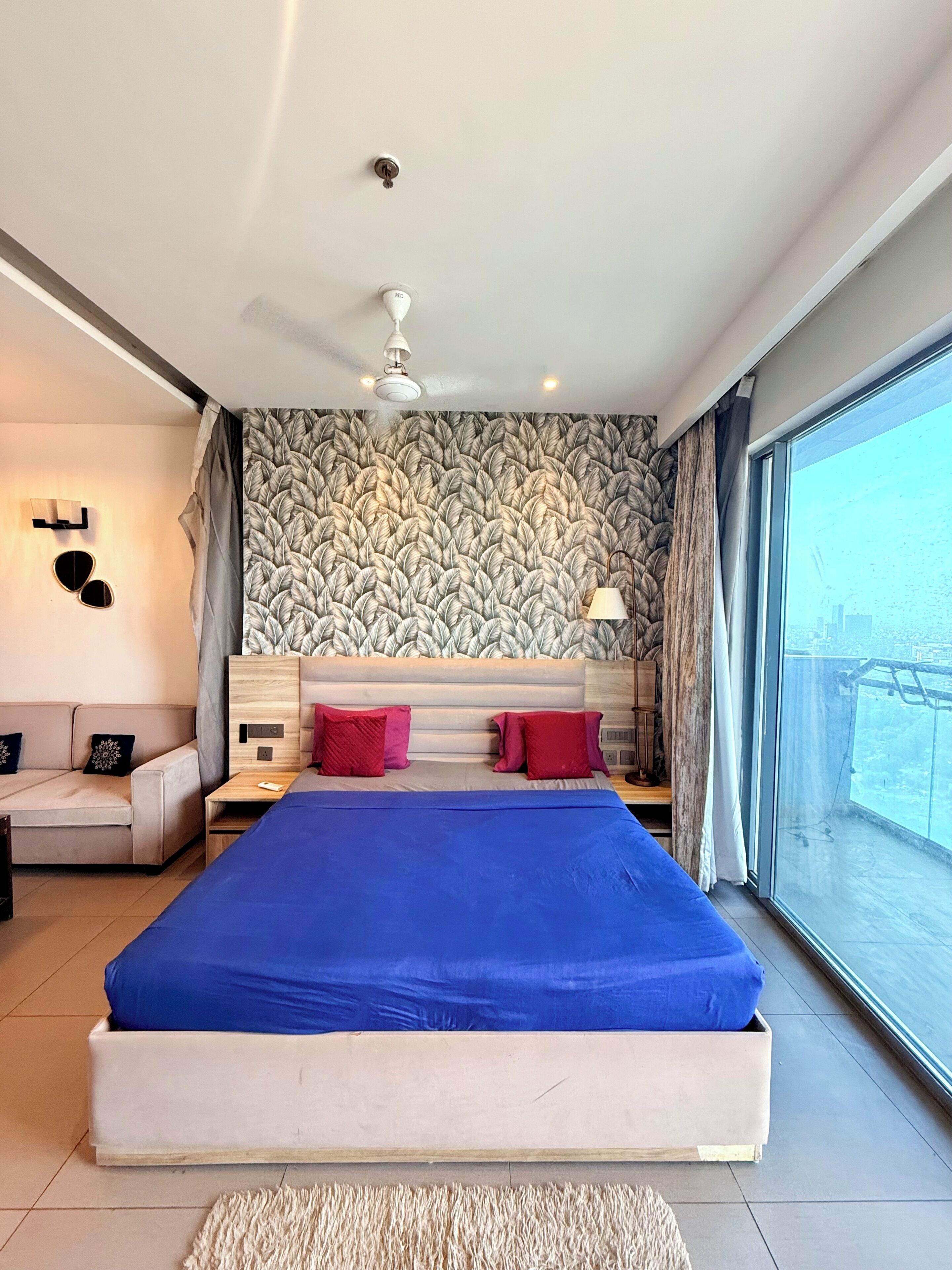 A modern bedroom with a large bed, a sofa, a ceiling fan, and a view of the city through large windows.