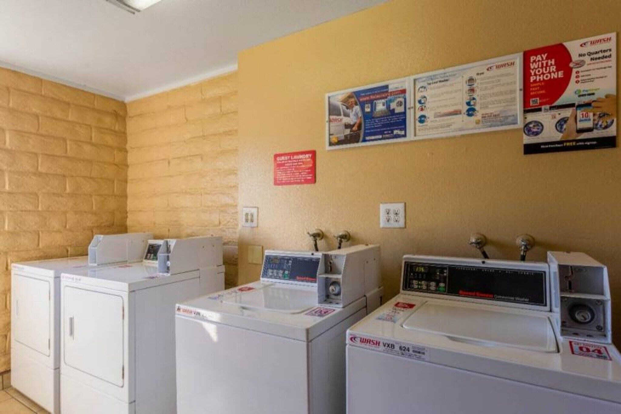 Un espace buanderie équipé de trois lave-linge et trois sèche-linge, un panneau indiquant qu’il s’agit de la buanderie pour les clients, et des affiches d’information accrochées au mur.