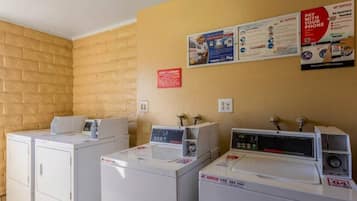 Un espace buanderie équipé de trois lave-linge et trois sèche-linge, un panneau indiquant qu’il s’agit de la buanderie pour les clients, et des affiches d’information accrochées au mur.