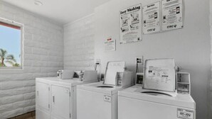 Ein Waschraum mit zwei Frontlader-Waschmaschinen, einem Trockner und einer an der Wand montierten Waschmaschine. An der Wand befinden sich Schilder mit den Aufschriften 'Easy Washing' und 'Service'.