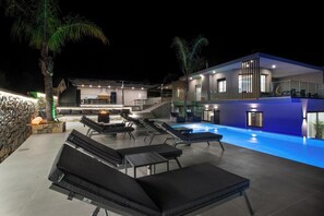 A modern outdoor pool area with lounge chairs, a stone wall, and a two-story building with a glass balcony. - Manel Villa - Mountain Whispers by etouri (Chania)