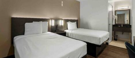 A hotel room with two beds, a wooden floor, a bathroom with a mirror and sink, and a small desk.
