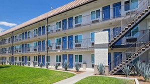 A multi-story motel with a central courtyard, featuring a row of rooms with blue doors, a staircase, and a well-maintained lawn.