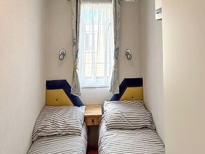 A small room with two beds, a window with curtains, and a wooden nightstand. - Horse Shoe Lakes - UKC9581 (Mablethorpe)