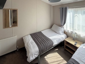 Ein Einzelbett mit einer grau-weißen Streifenauflage, ein Nachttisch mit Lampe und ein Fenster mit Vorhängen.