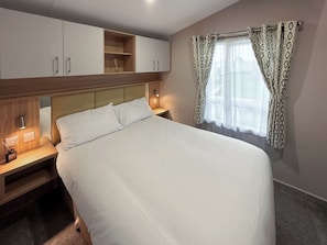 Ein ordentlich bezogenes Bett in einem Hotelzimmer mit Kopfteil, Nachttischen und einem Fenster mit gemusterten Vorhängen.