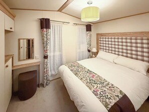 A bedroom with a bed, a bedside table with a lamp, a mirror, and curtains. - Oak Village 83 - UKC9580 (Mablethorpe)