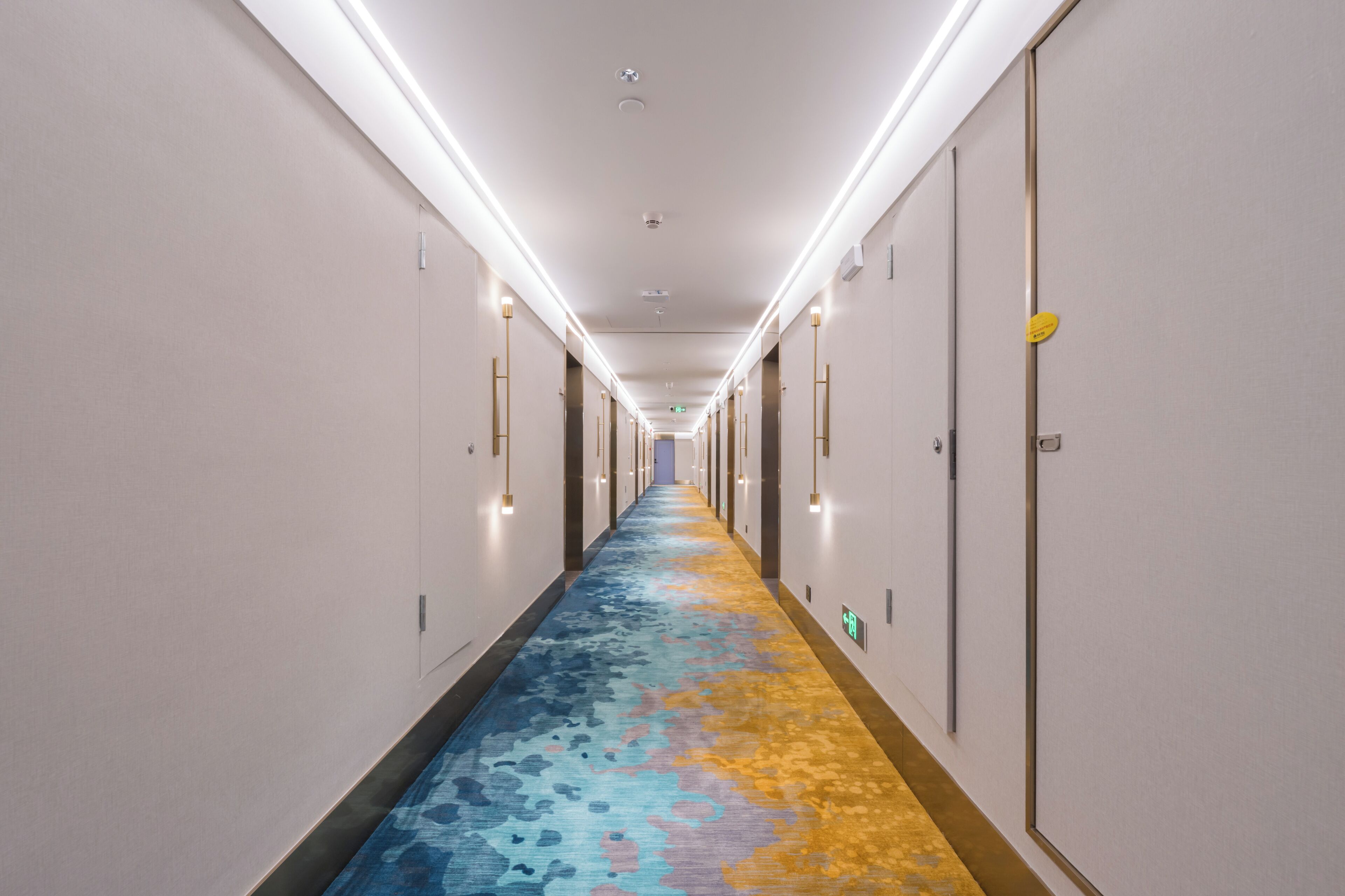 A hotel hallway with a patterned carpet, doors on both sides, and overhead lighting.