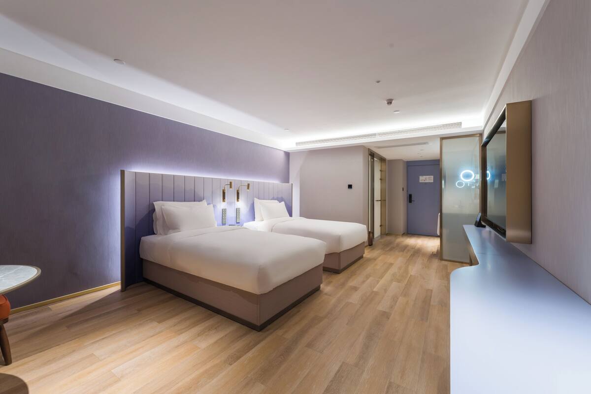 A modern hotel room with two beds, a small table, a mirror, and a bathroom visible through an open door.