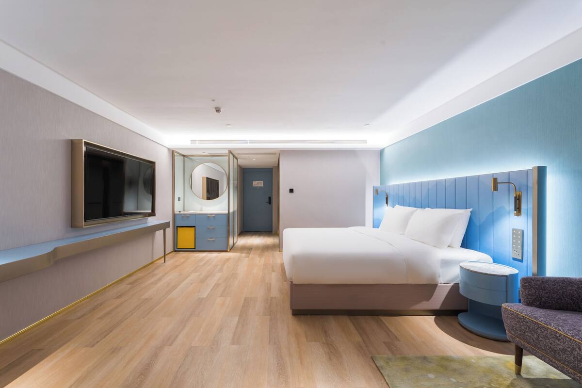 A modern hotel room with a large bed, a television, a bathroom with a mirror, and a blue accent wall.