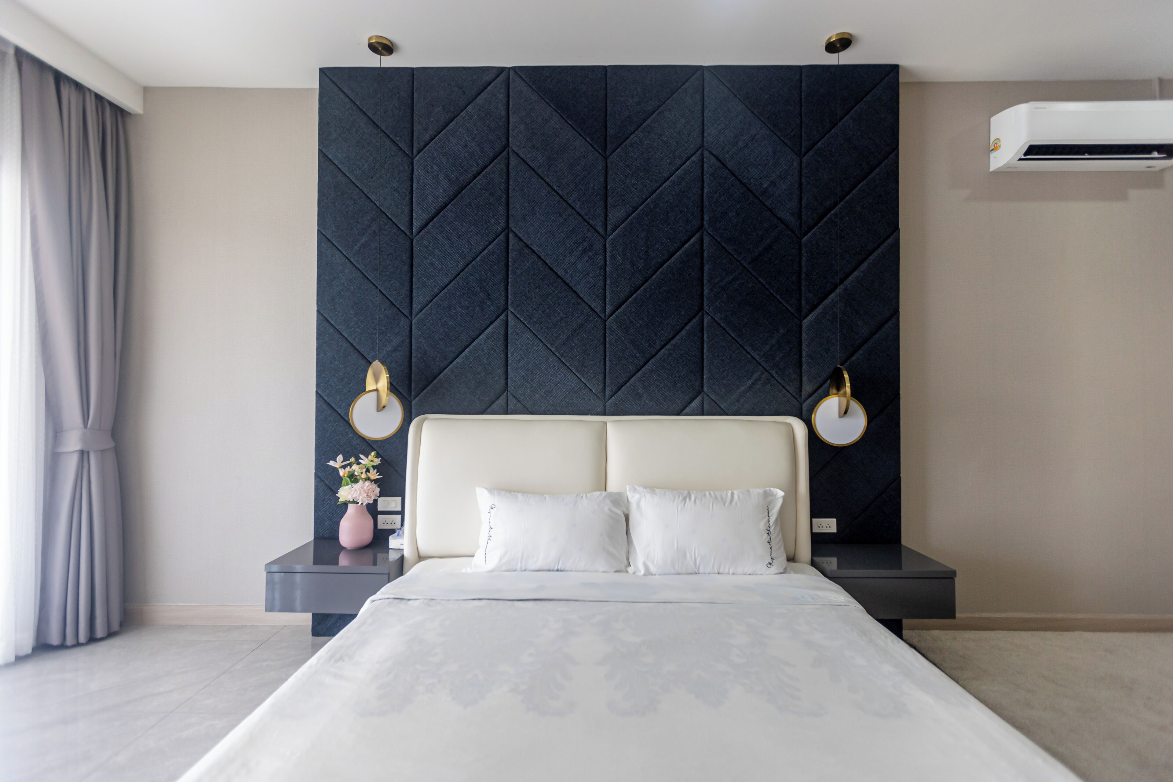 A modern hotel room with a large bed, a dark textured headboard, bedside tables with lamps, and an air conditioning unit on the wall.