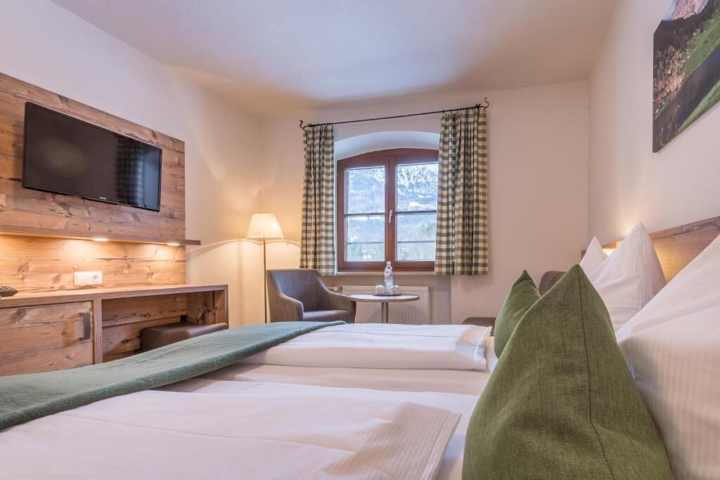 A hotel room with a bed, a TV mounted on a wooden panel, a chair, a small table, and a window with curtains.