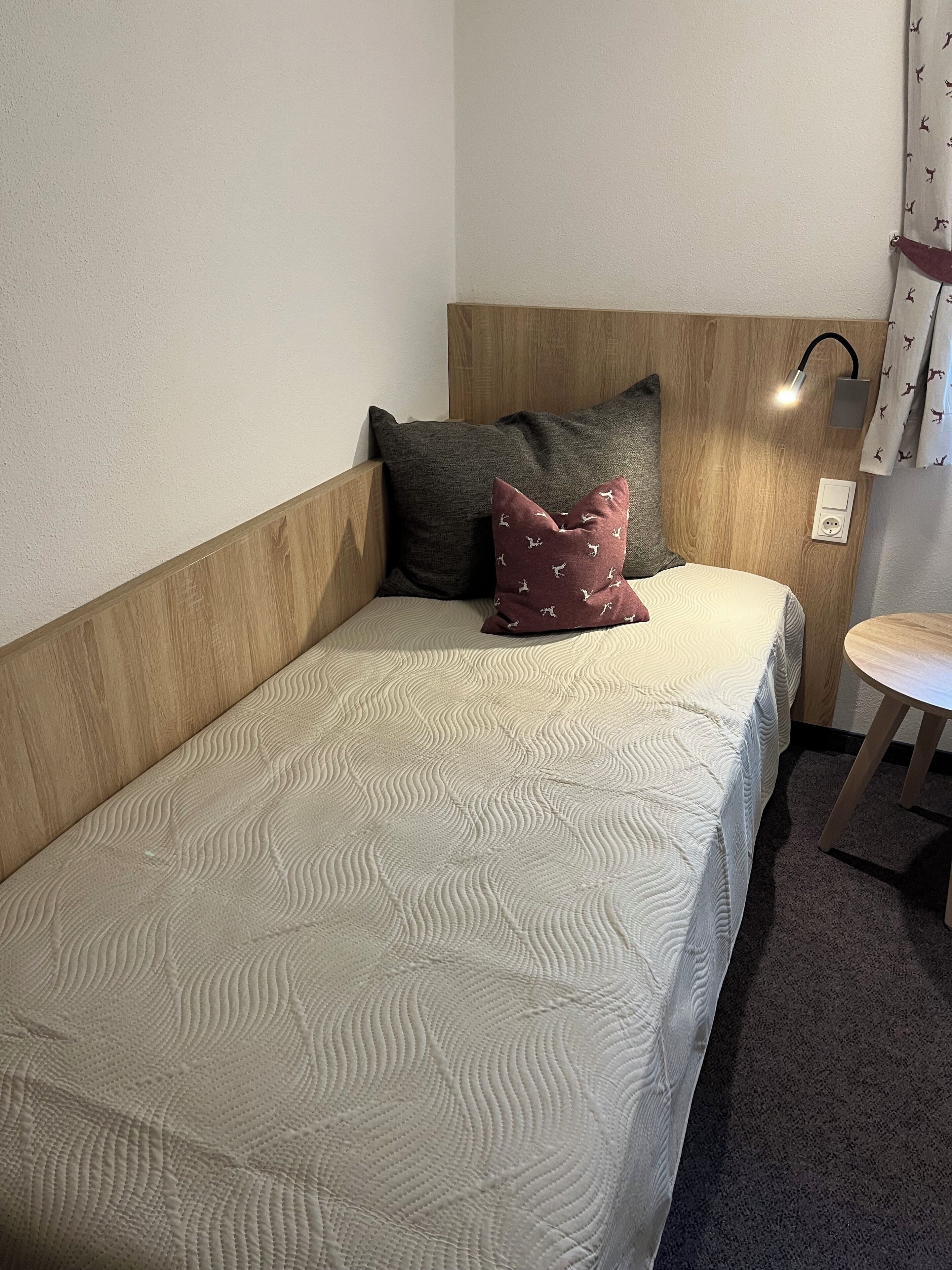 Una cama individual con una colcha blanca texturizada, dos almohadas, una mesita de noche y una lámpara de pared.