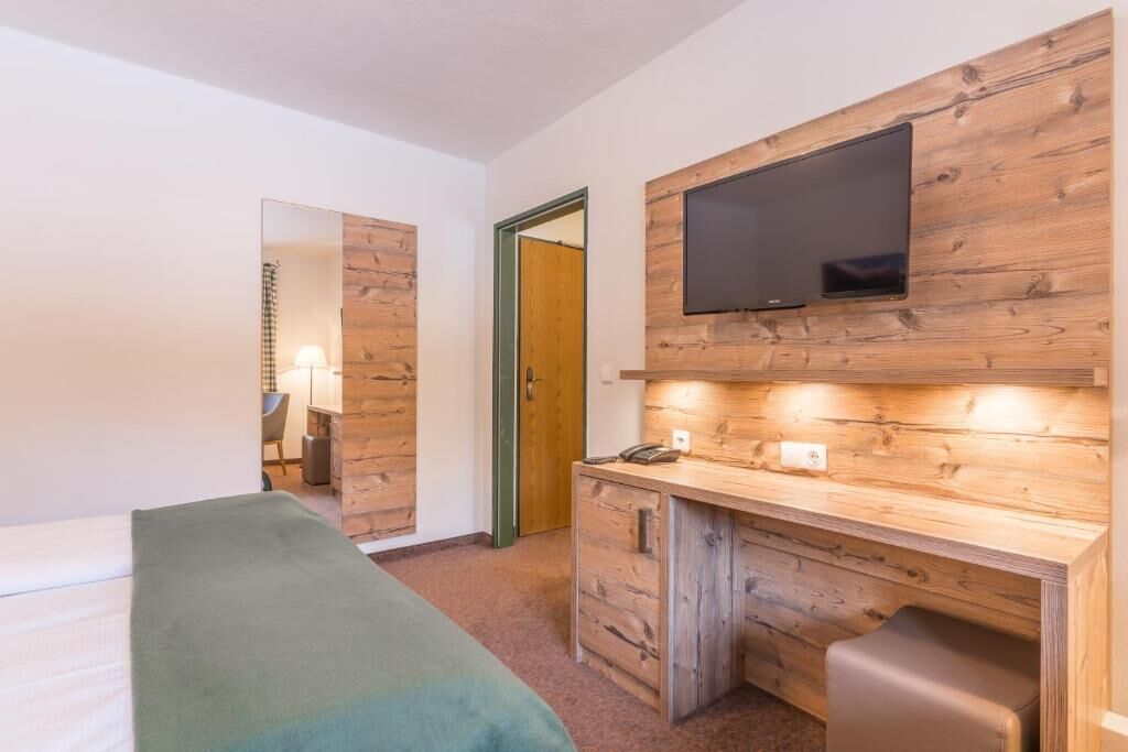A room with a bed, a wooden desk, a television mounted on the wall, and a mirror.