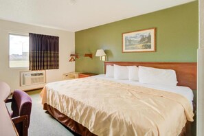 A hotel room with a large bed, a desk, a chair, a window with curtains, an air conditioning unit, and a painting on the wall.