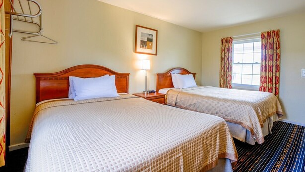 A hotel room with two beds, a bedside table with a lamp, a framed picture on the wall, and a window with curtains.