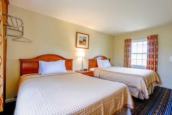 A hotel room with two beds, a bedside table with a lamp, a framed picture on the wall, and a window with curtains.