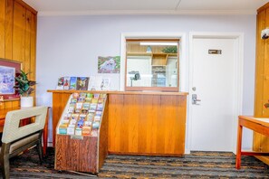 Un comptoir d’accueil avec un plan de travail en bois, un panneau d’affichage où sont accrochées des brochures de voyage, un tableau encadré au mur et une porte avec une pancarte.