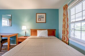 Una habitación de hotel con cama, escritorio, lámpara, un cuadro enmarco y una ventana con cortinas.