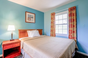 A hotel room with a bed, a nightstand, a lamp, a window with curtains, and a framed picture on the wall.