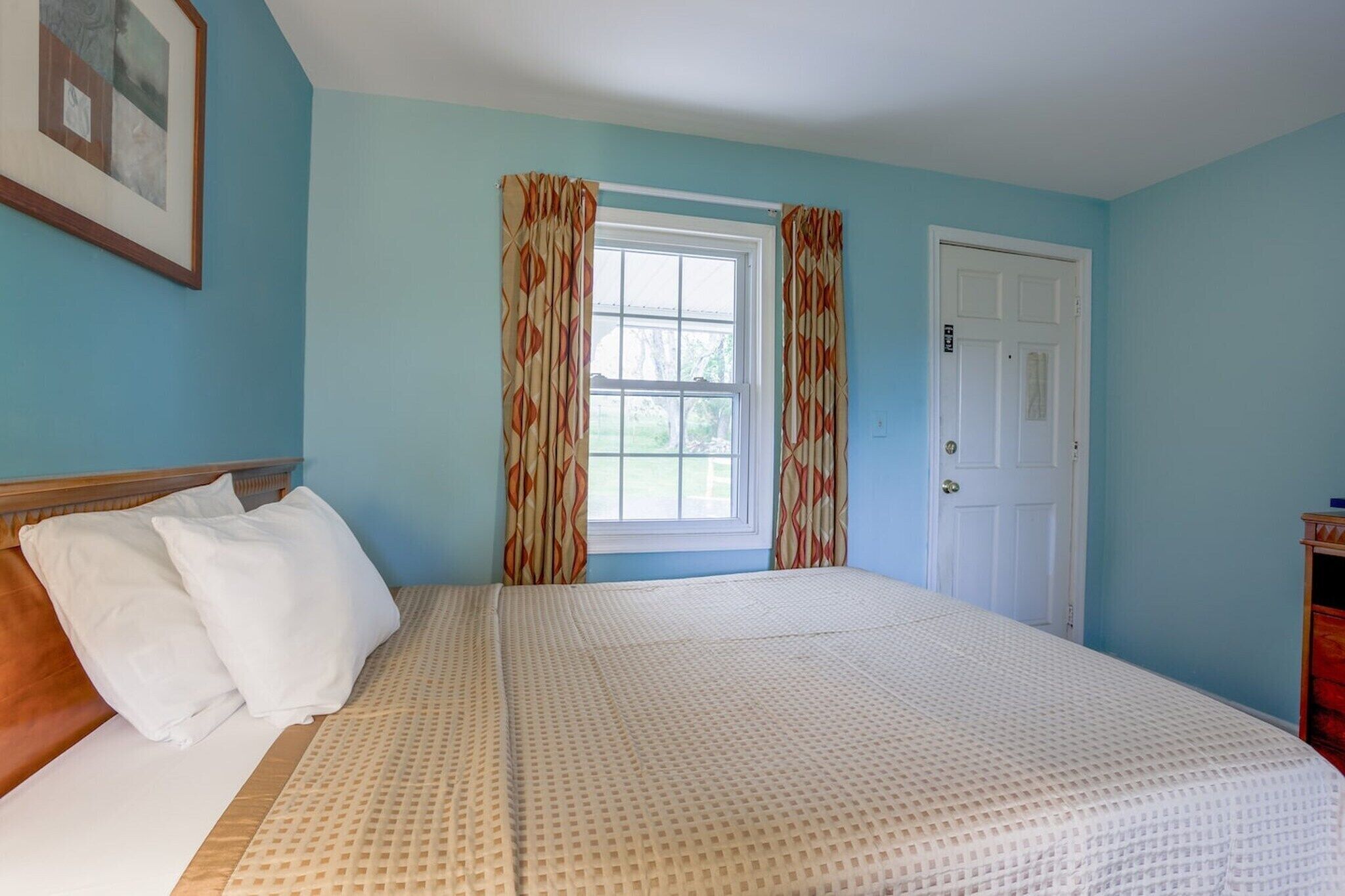 A bedroom with a bed, a window with curtains, a door, and a framed picture on the wall.