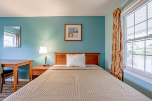 A hotel room with a bed, a desk, a lamp, a framed picture, and a window with curtains.
