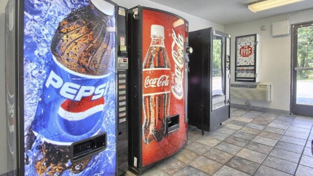 Pepsi- ja Coca-Cola-automaatit käytävällä, jossa on laatoitettu lattia ja oikealla puolella ovi.