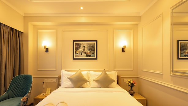 Habitación de hotel con cama, dos lámparas de pared, un cuadro enmarcado, una silla, una mesita de noche y un jarrón con flores.